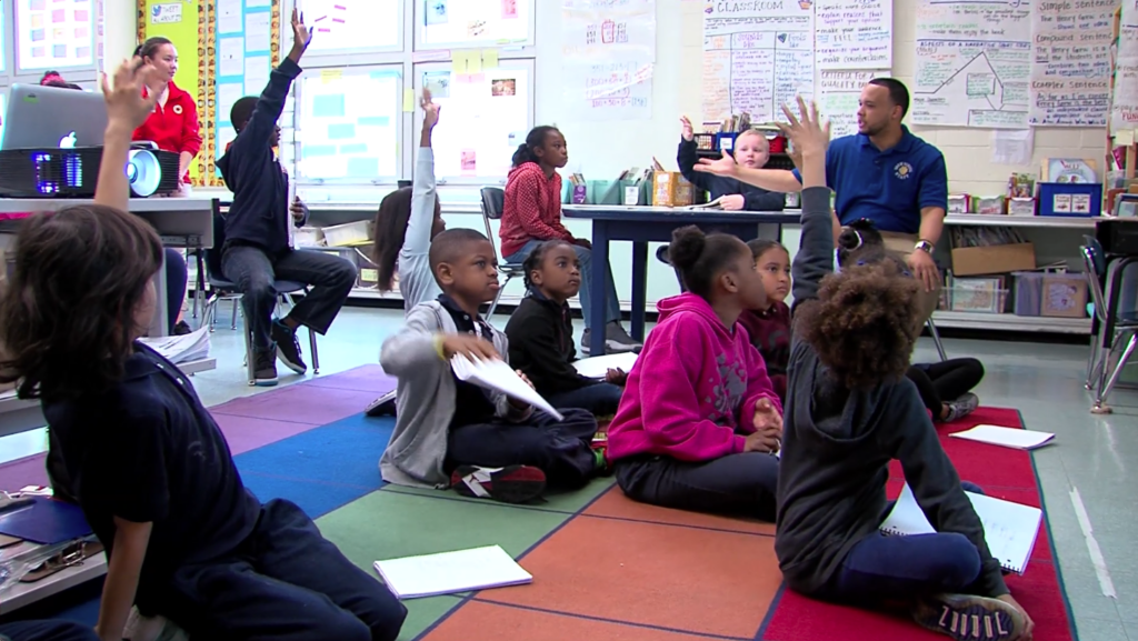 A third grade teacher facilitates a whole class discussion with a diverse group of students. He sits at a table with two children. The rest of the students are on the rug or in chairs. Many have their hands raised.